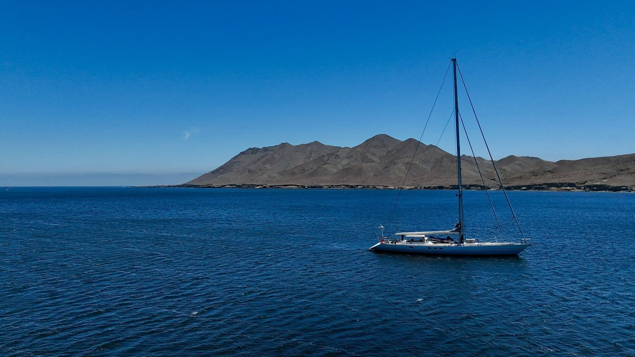 A sailboat on calm blue waters with a mountainous island in the background under a clear blue sky.