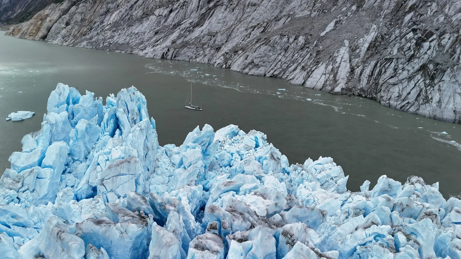 Blue icebergs floating near a rocky shoreline with a sailboat in the water