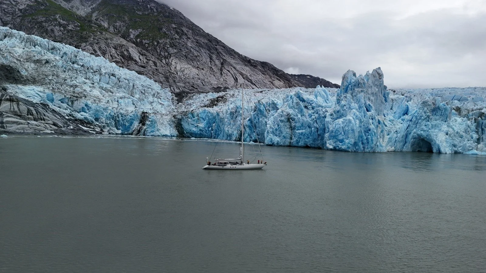 A sailboat floating on calm water near a large glacier with ice and rock formations in the background.