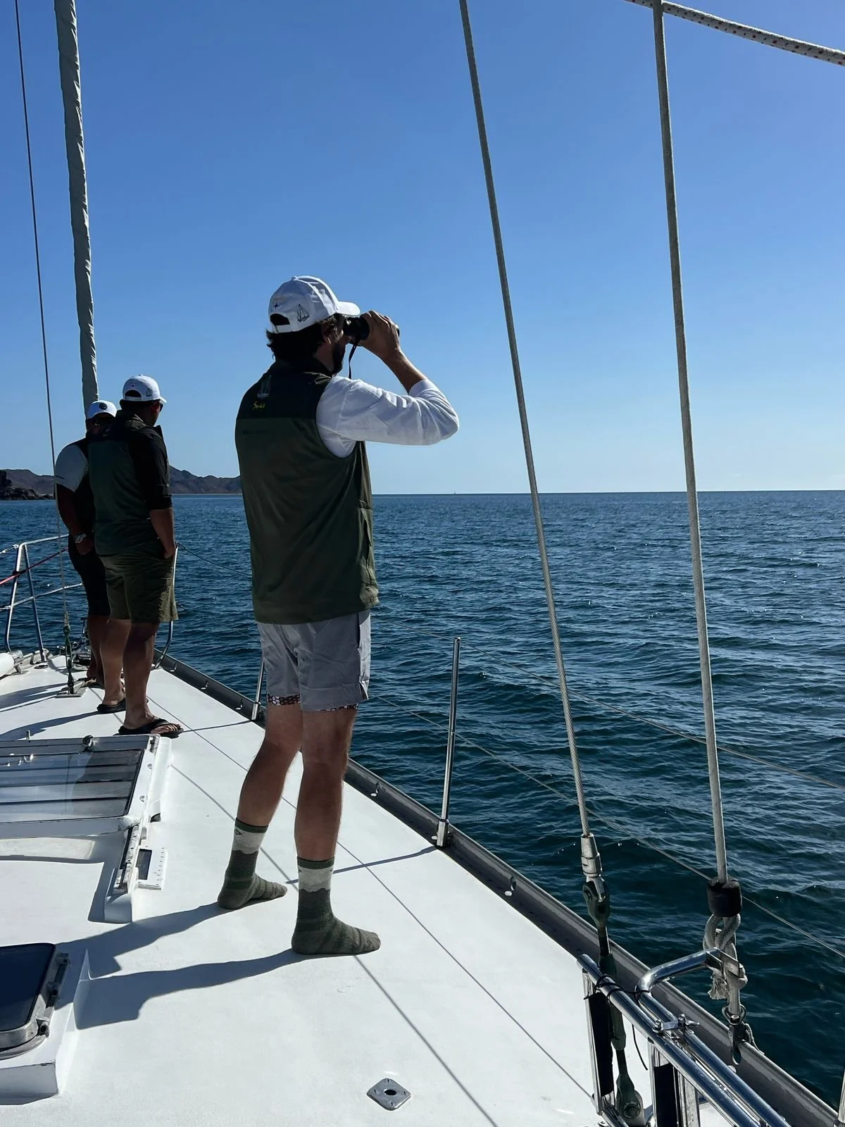 Three men standing on a sailboat deck looking out at the ocean under a clear blue sky, one of whom is using binoculars.