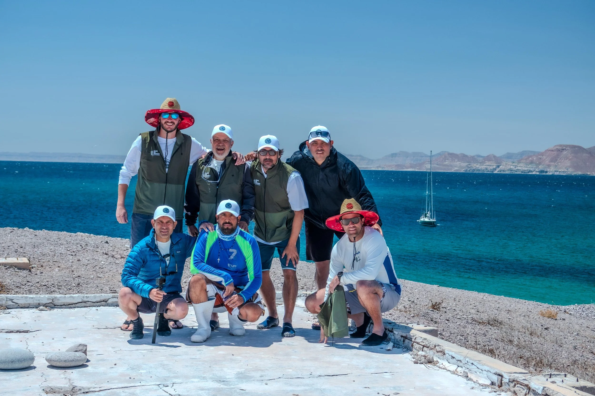 Group of eight men posing outdoors near a lake with mountains in the background. They are dressed in casual and outdoor clothing, some wearing hats and sunglasses. One man is holding a walking stick. The weather is sunny, and a sailboat is visible on