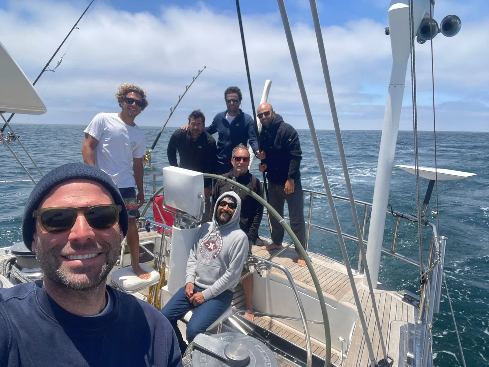 Group of seven men on a sailboat in the ocean during daytime, taking a selfie, with some holding fishing rods.