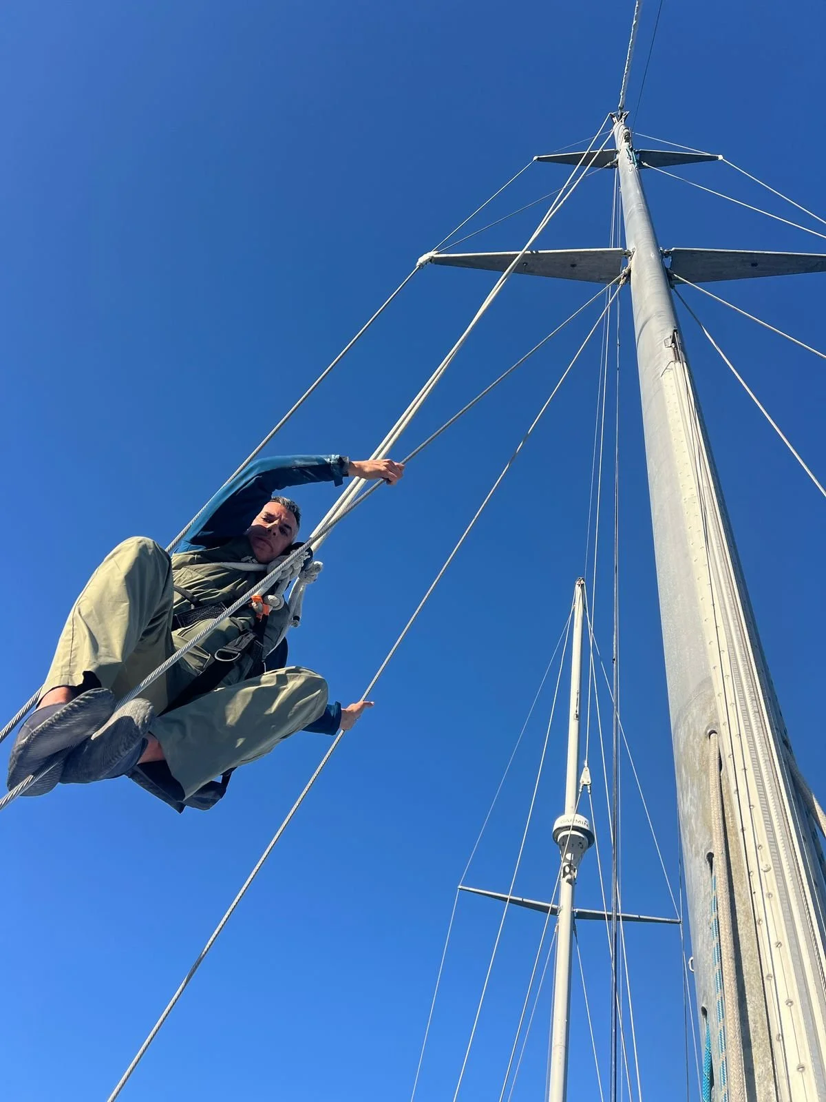 Person climbing the mast of a sailboat against a clear blue sky.