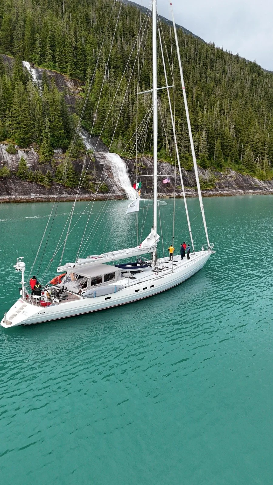 A white sailboat on a turquoise lake with a forested mountain and waterfall in the background. Several people are on the boat, some standing and some sitting.