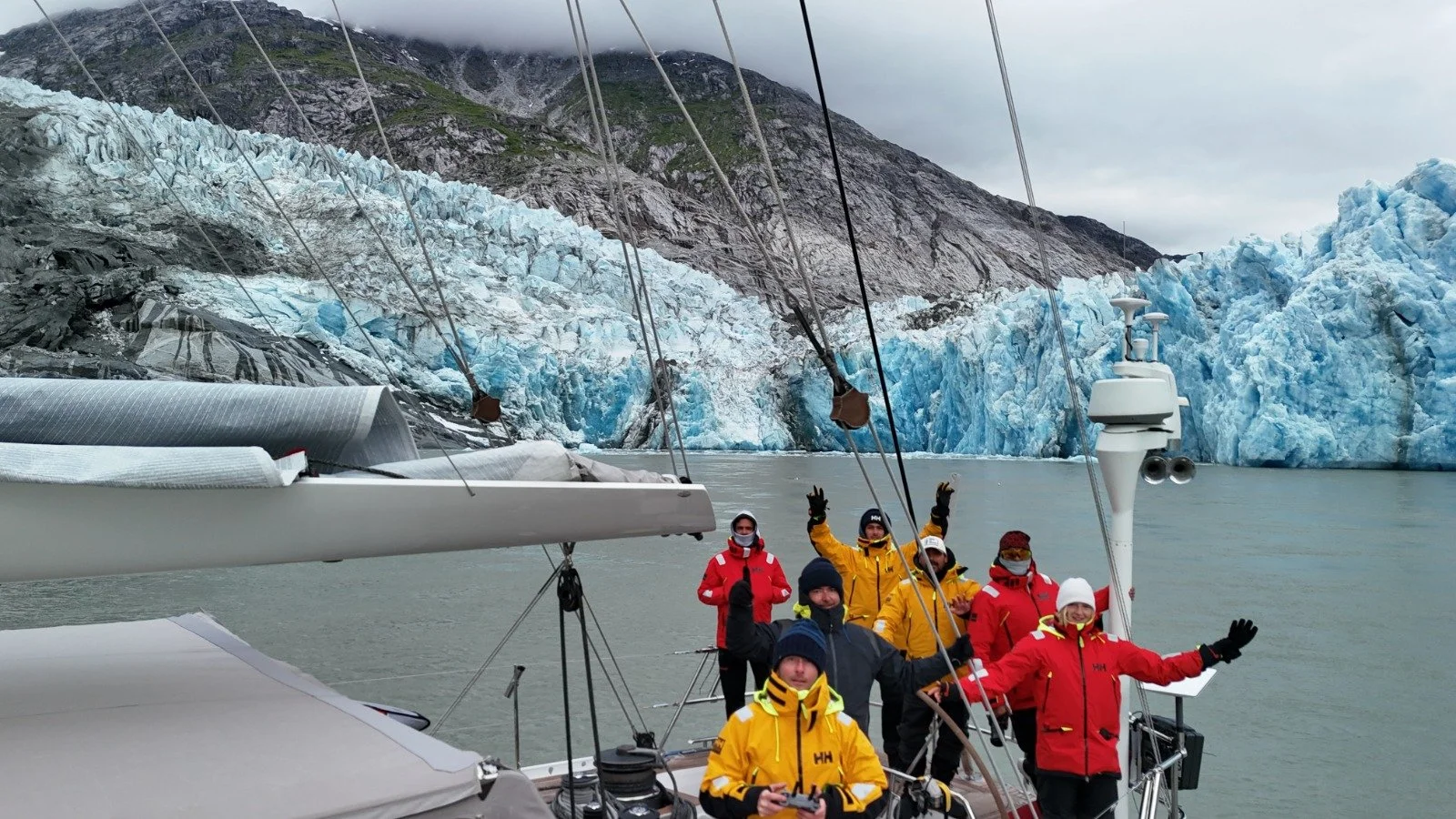 Group of people on a sailboat near a glacier with mountainous terrain in the background.