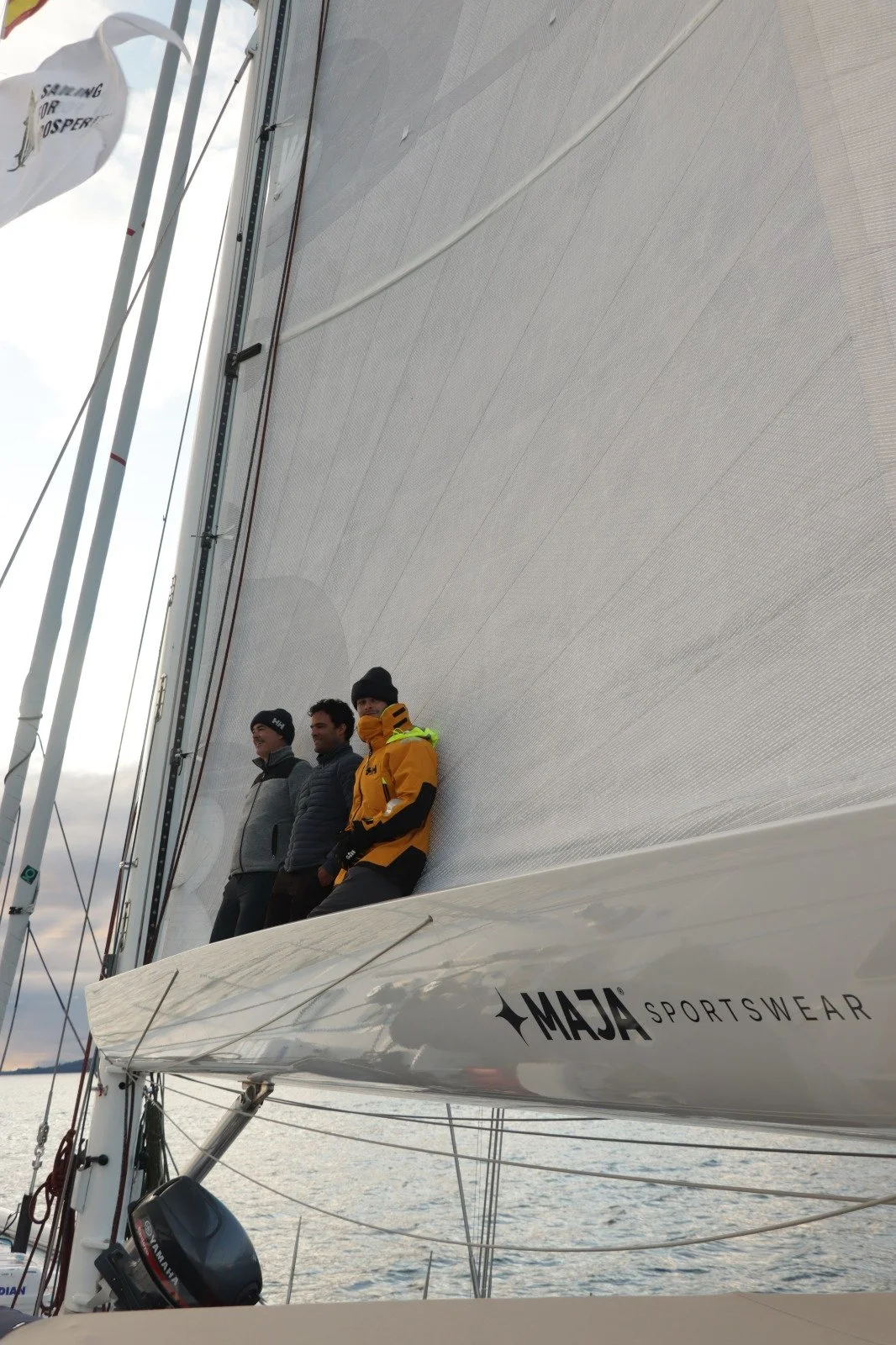 Three men standing on the edge of a sailboat's hull by the water, with large sail raised overhead and cloudy sky background.