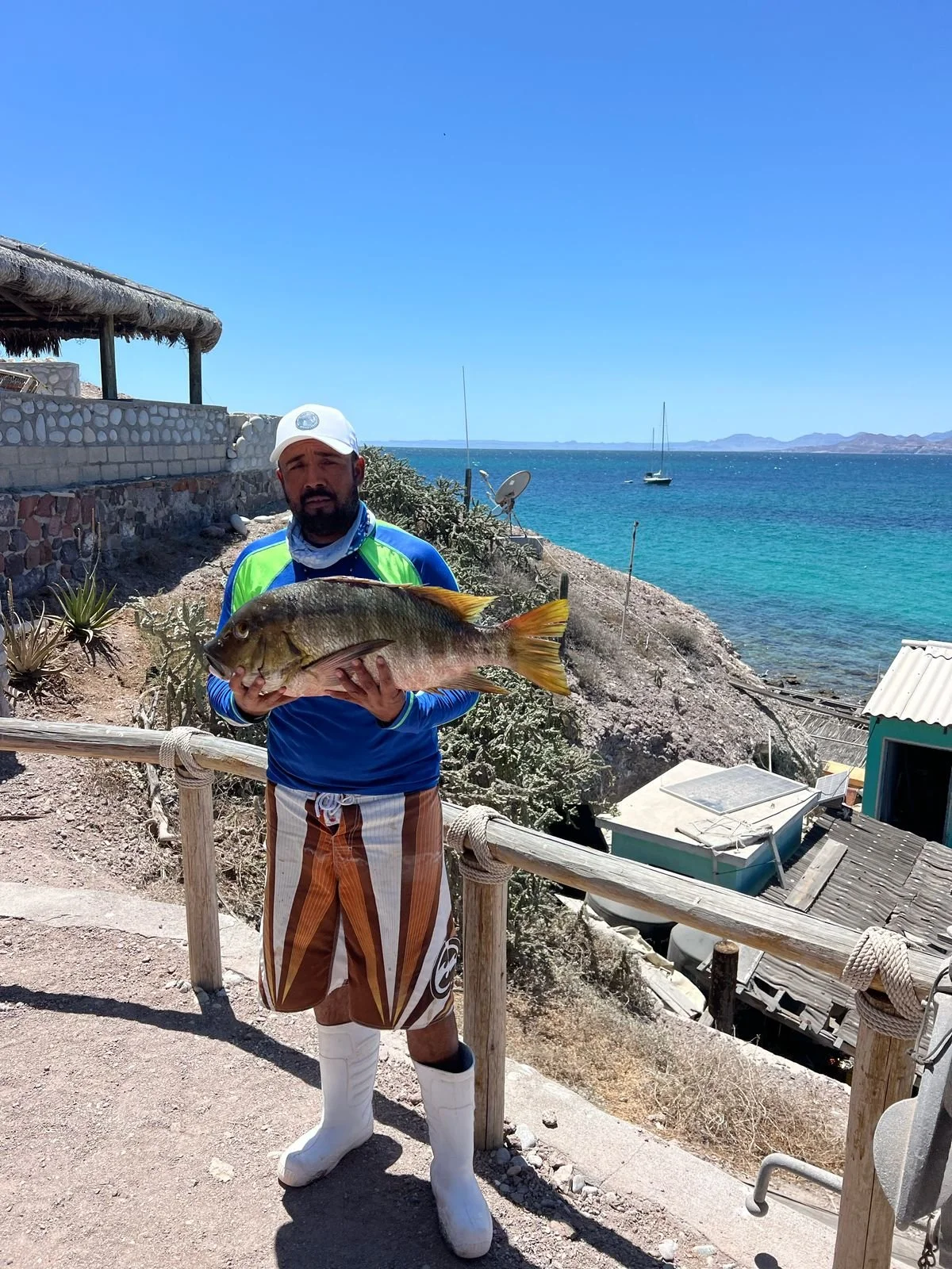 Man holding a large fish by the coastline with the ocean and sailboats in the background.