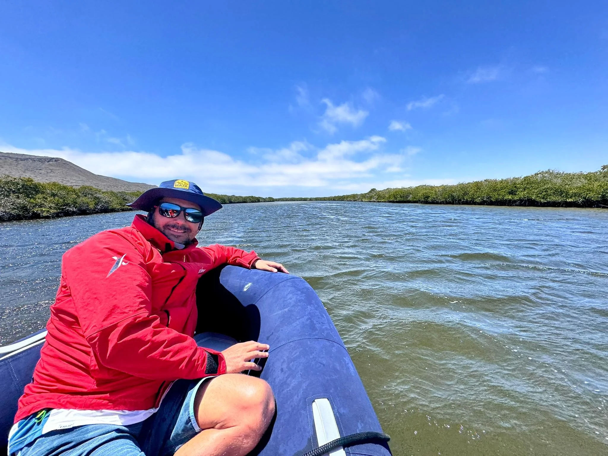 Man in red jacket, sunglasses, and wide-brimmed hat sitting in inflatable boat on a river with trees on both sides and blue sky with clouds.