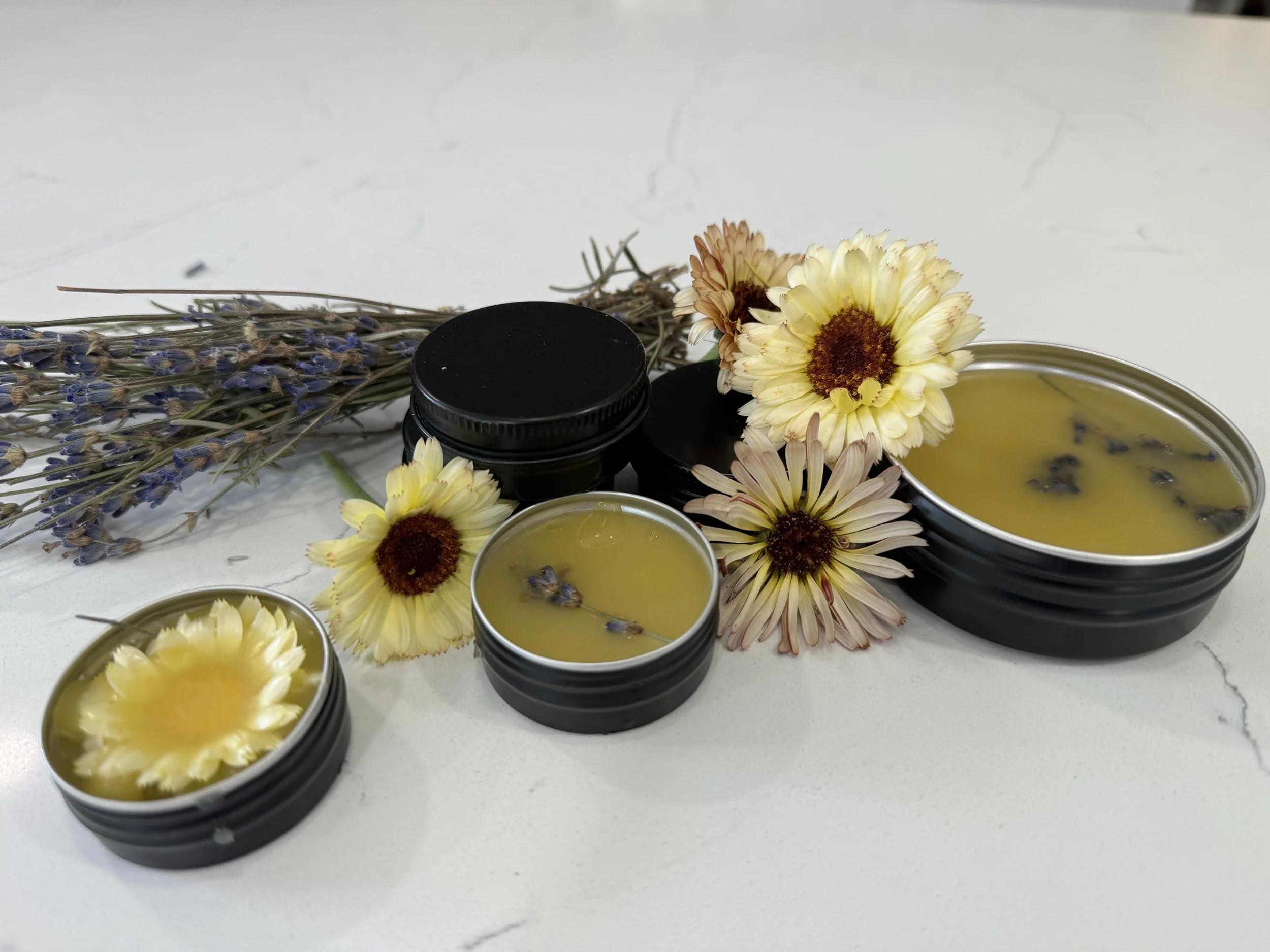 Black containers filled with calendula salve, surrounded by dried lavender and flowers, on a white marble surface.