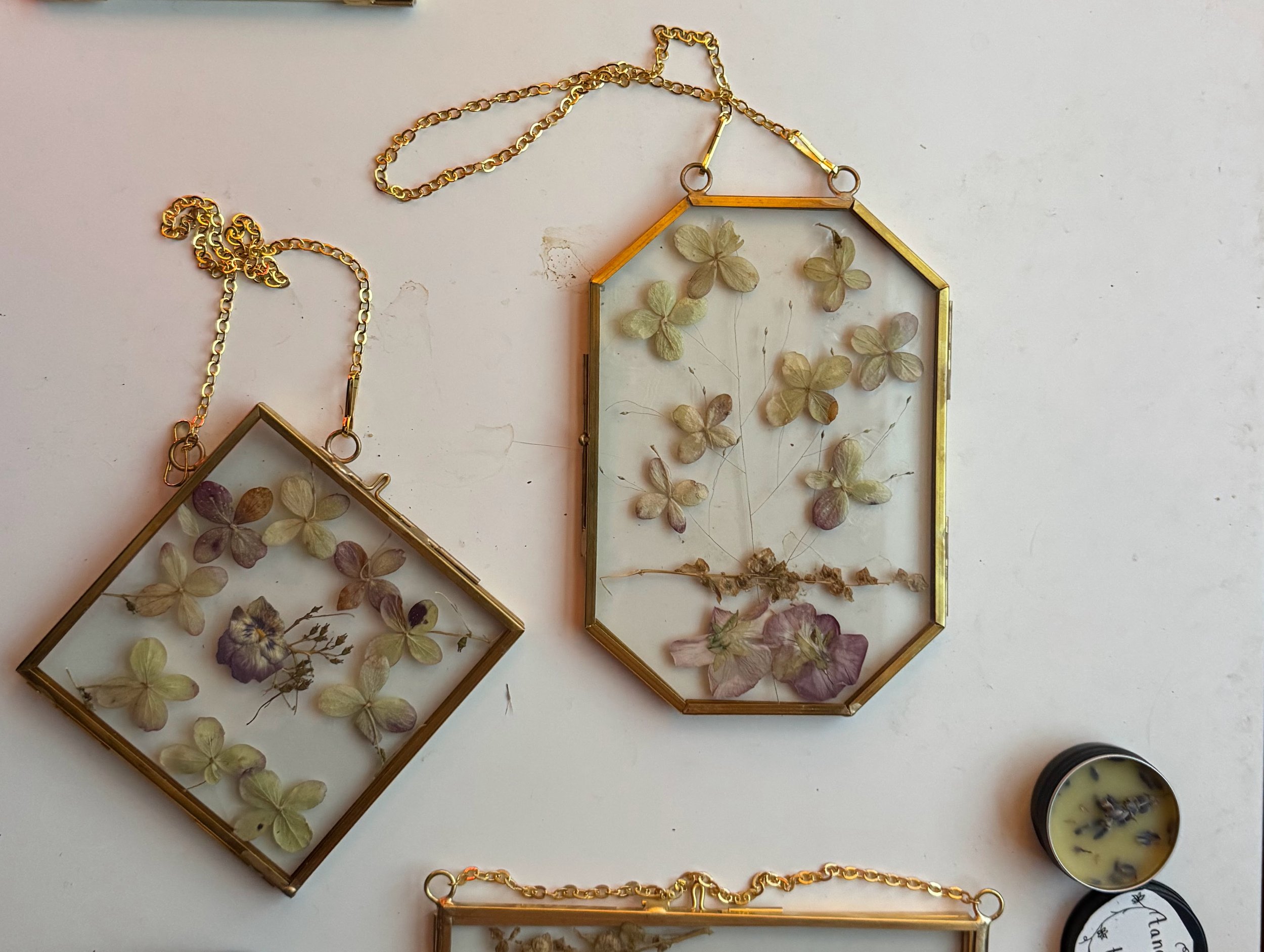 Two decorative glass frames with pressed dried flowers and butterflies, hanging from gold chains on a white surface, and a small black container with homemade salve on the right side.