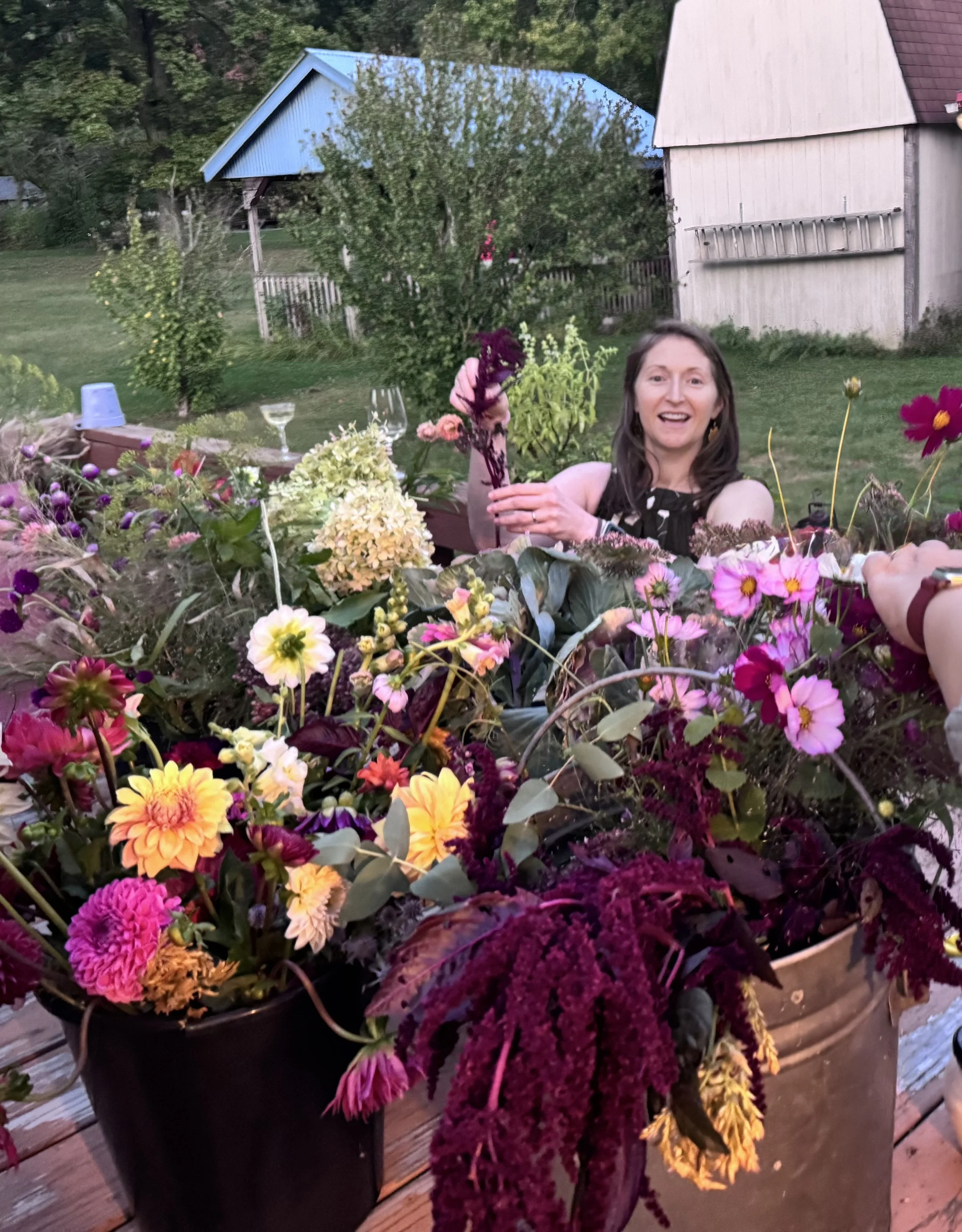 A woman with long brown hair smiling and holding a purple flower, sitting at a table adorned with a variety of colorful flowers in pots, outdoors in a backyard with trees, a fence, and two houses with blue and brown roofs in the background.