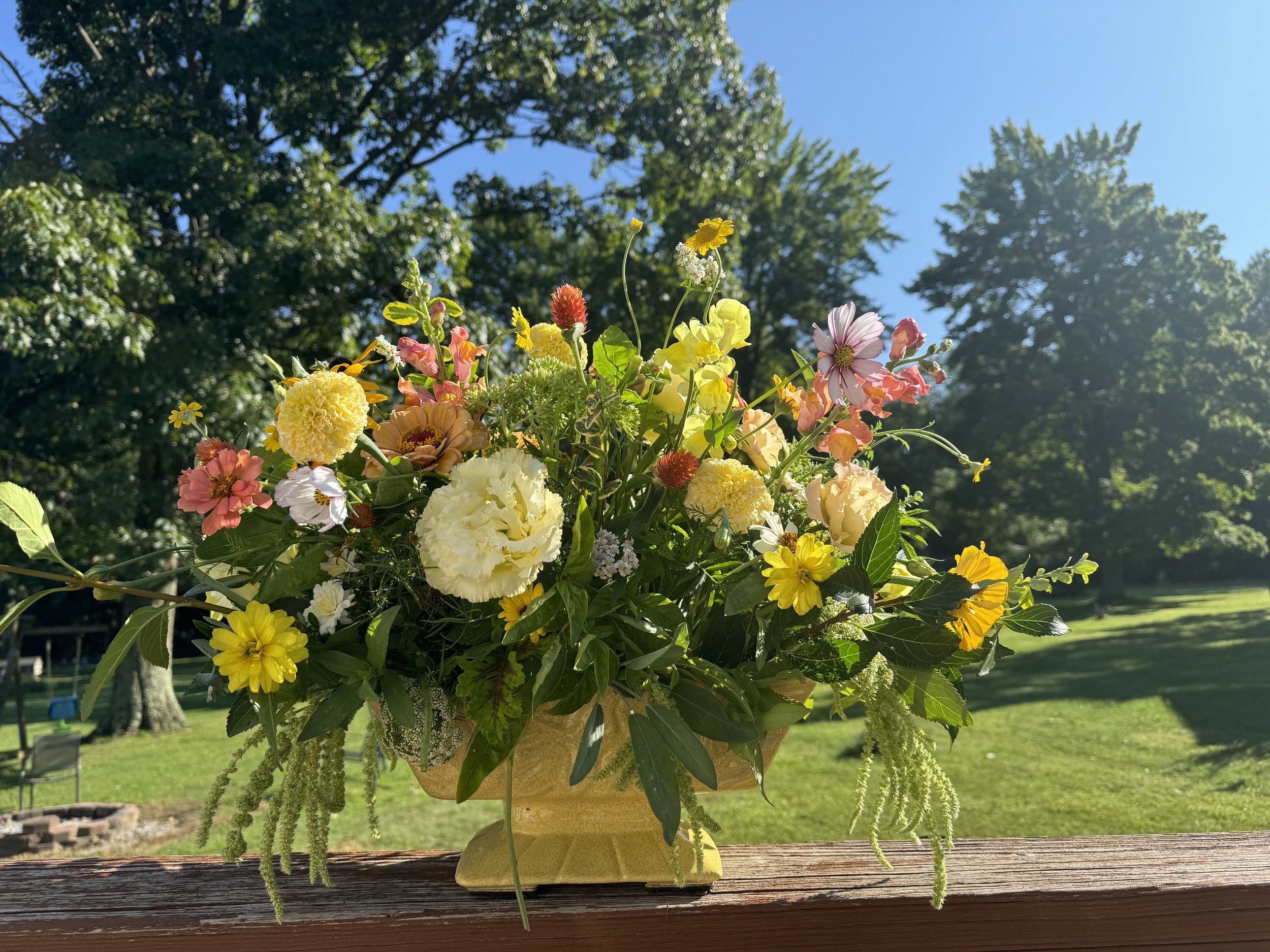 A large yellow ceramic vase filled with a variety of colorful fresh flowers including dahlias,marigolds, daisies, roses, and other flowers, placed on a wooden railing outdoors on a sunny day with green trees and a blue sky in the background.
