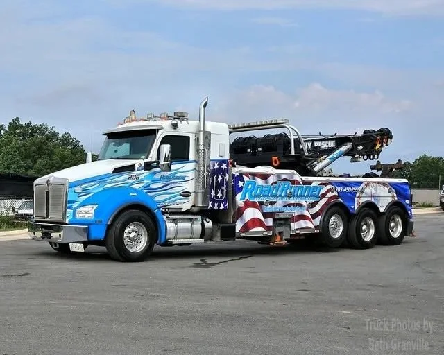 A road rescue tow truck decorated with patriotic American flag graphics, parked on a paved surface under a partly cloudy sky.