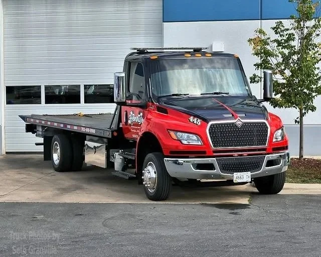 Red and black flatbed tow truck parked in front of a building with a white and blue wall.