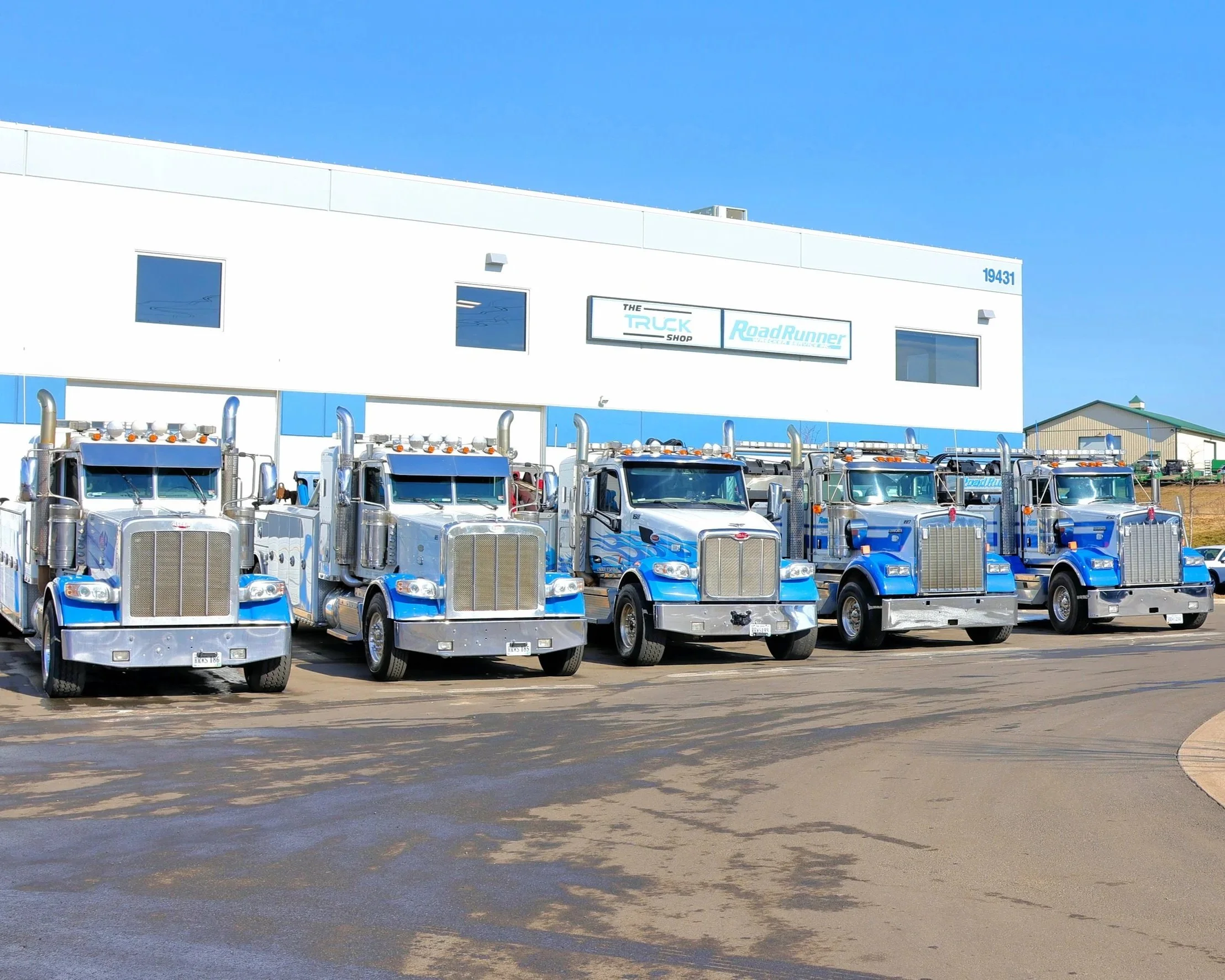 Five blue and silver semi trucks parked in front of a white building with signs that read 'The Truck Shop' and 'RoadRunner' during daytime.