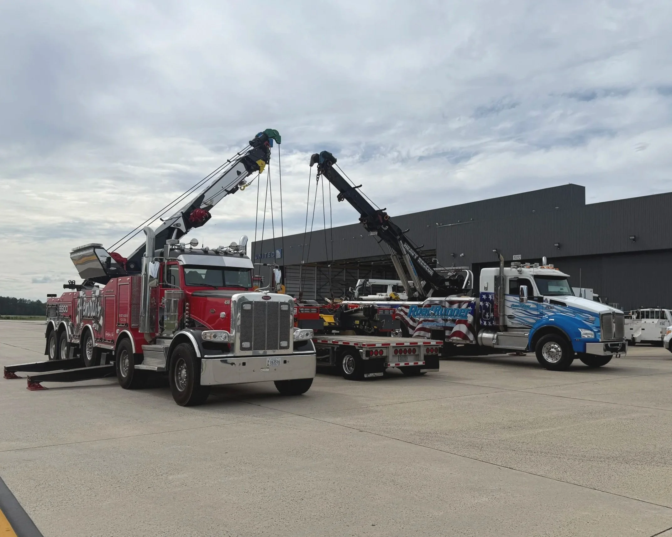 Two large tow trucks with cranes are parked on an airport tarmac, with a large hangar in the background and overcast skies above.