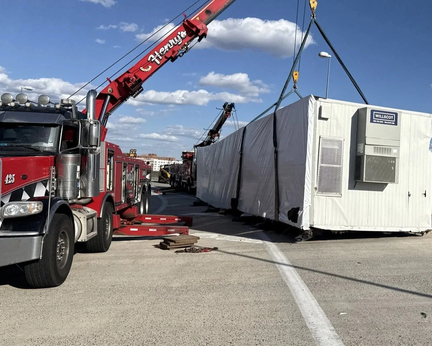 Emergency response scene with a large white temporary building being lifted by a red tow truck crane on a highway under a cloudy sky.