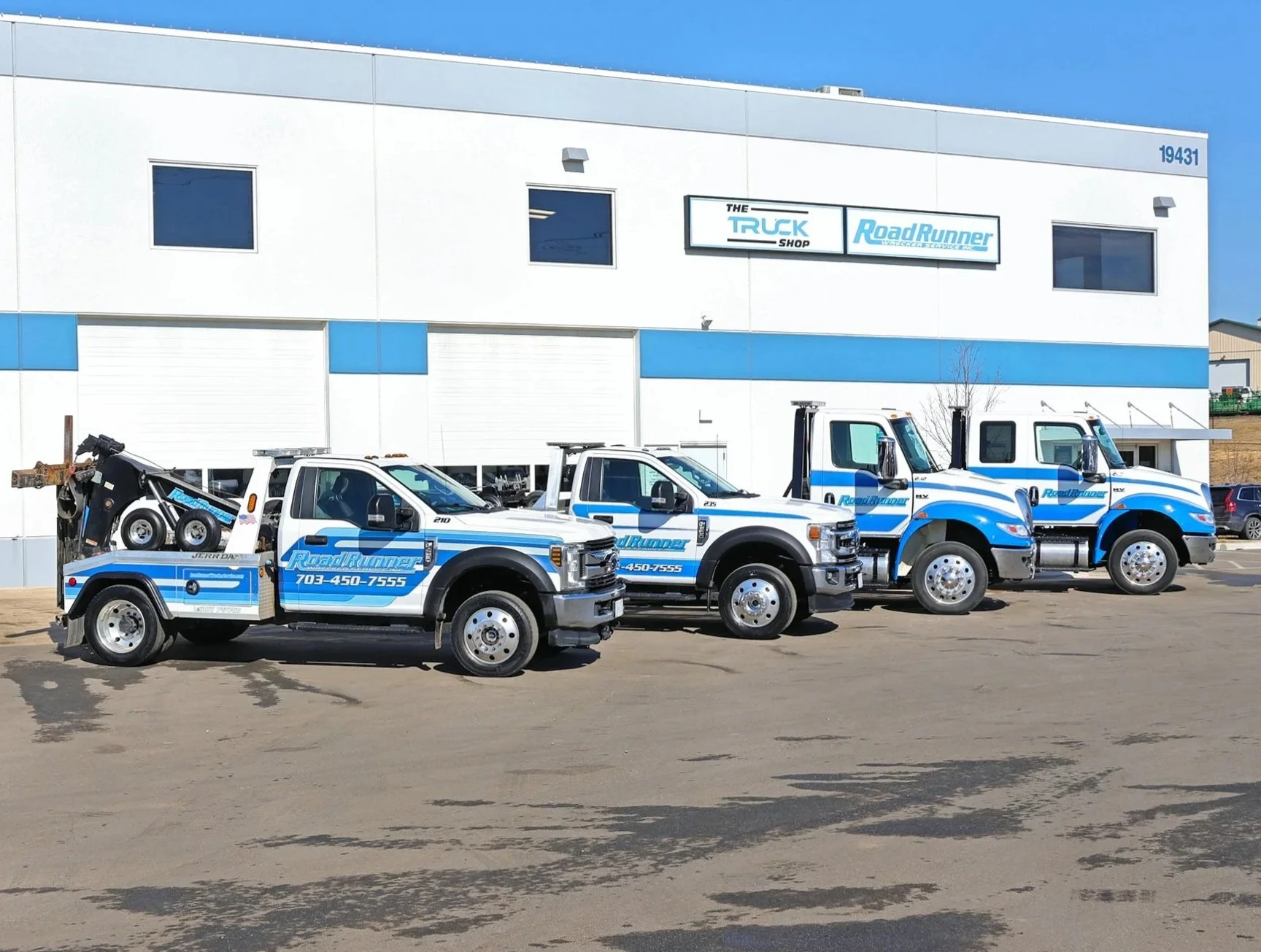 Four tow trucks parked in front of a white and blue building with signs reading 'The Truck Shop' and 'Road Runner.' The trucks have blue and white branding and are parked on an asphalt lot.