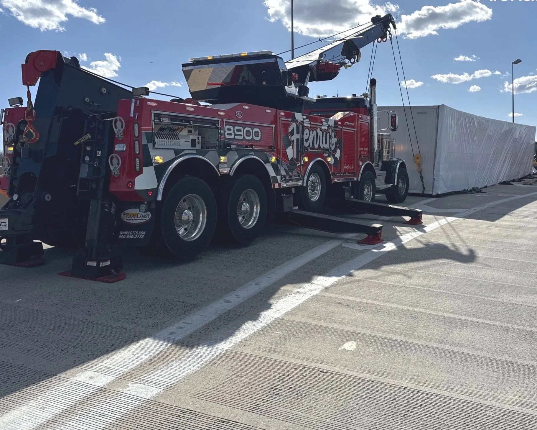 A red and black mobile crane with the label 'Henaut 8900' lifting a large, wrapped object on a rooftop parking lot under a partly cloudy sky.