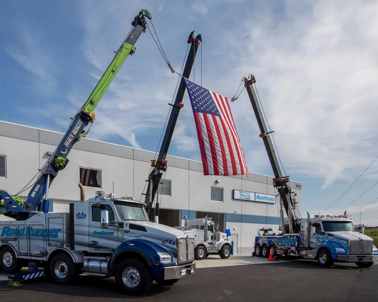 Two large tow trucks with extended arms lifting an American flag outside a commercial building under a partly cloudy sky.