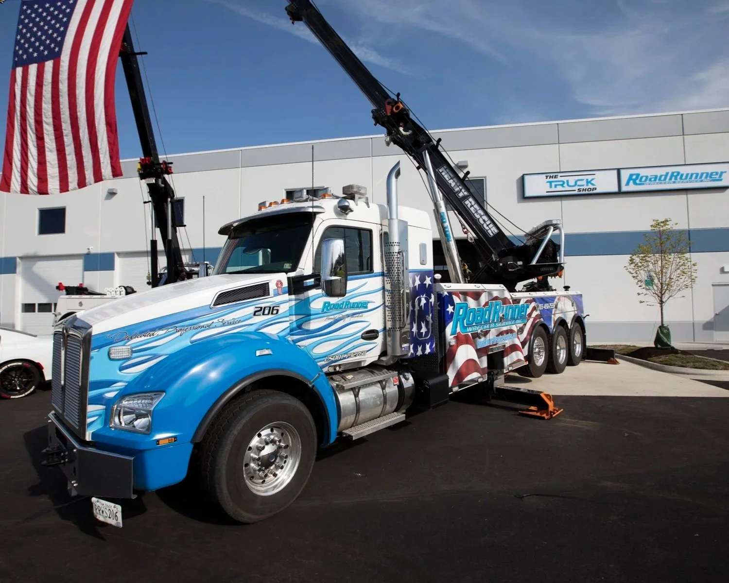 A tow truck decorated with an American flag design, parked outside a building with a sign that reads 'The Truck Shop Road Runners.' The truck is lifting a large American flag on a crane.