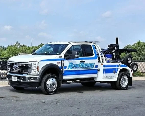 A white Ford utility truck with blue and gray stripe accents and the words "Road Runner" on the door, parked on an asphalt lot with a black fence and green trees in the background.