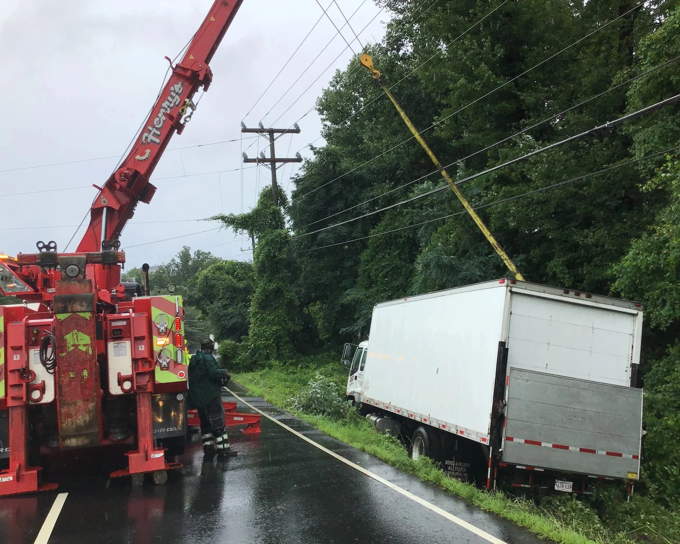 A truck crashed into power lines on a rainy road, with firefighters on scene using a crane to lift the fallen lines.