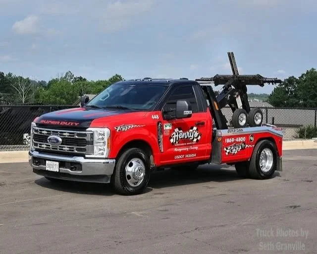 Red tow truck with Henry's Emergency Company logo, black hood, and towing equipment parked on a paved lot.