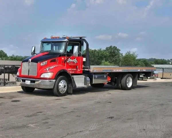 Red flatbed tow truck parked outdoors on asphalt with cloudy sky and trees in the background.
