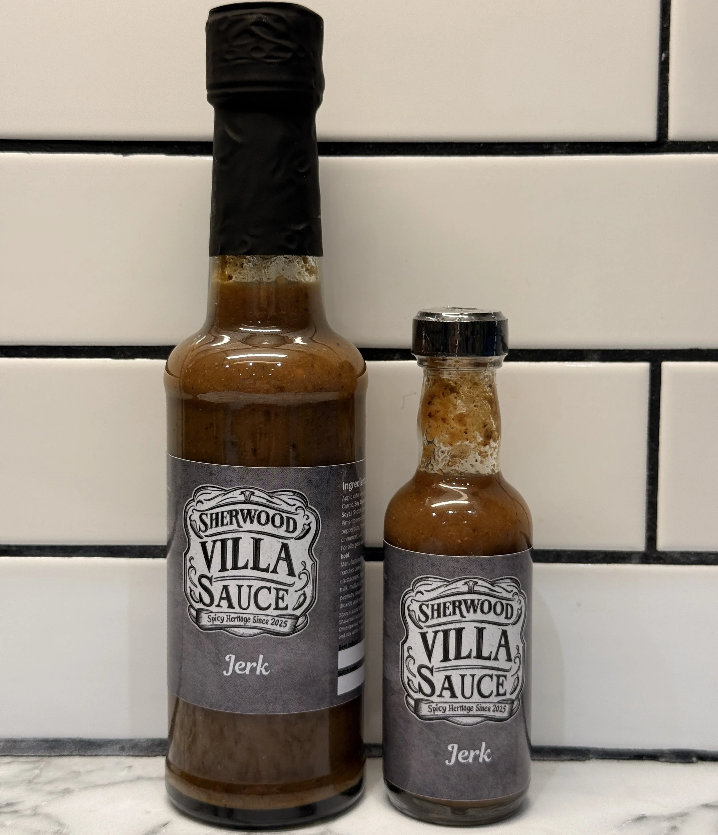 Two bottles of Sherwood Villa Sauce Jerk sitting on a white marble countertop against a white subway tile backsplash.