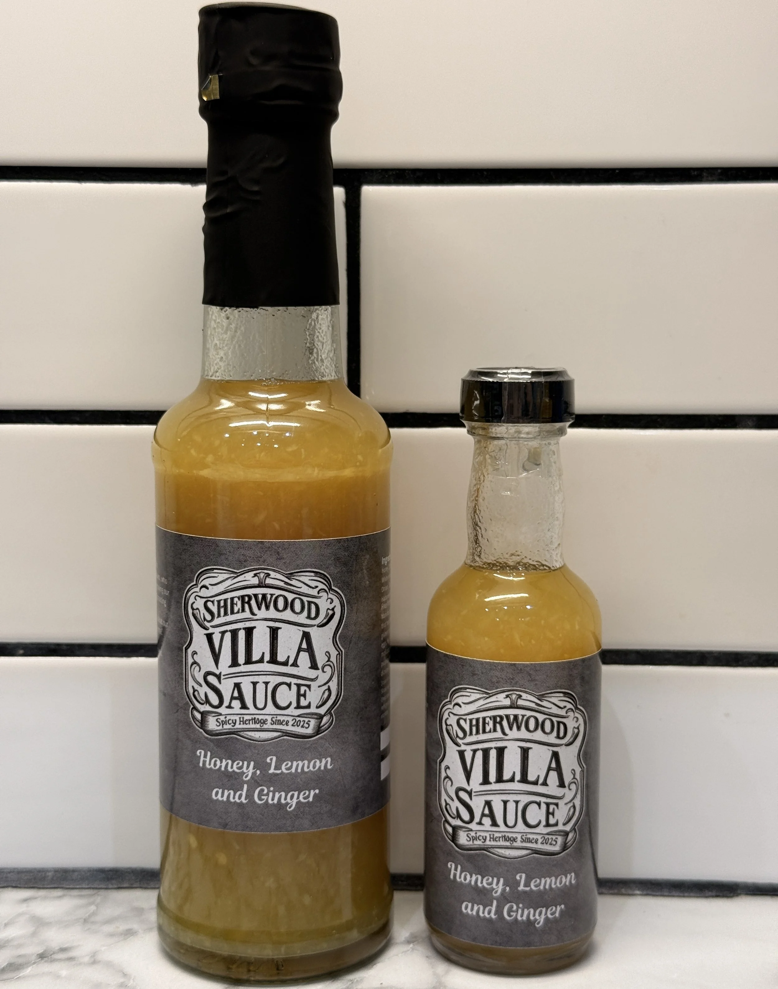 Two bottles of Sherwood Villa Sauce honey, lemon, and ginger in different sizes on a marble countertop in front of a white tiled wall.