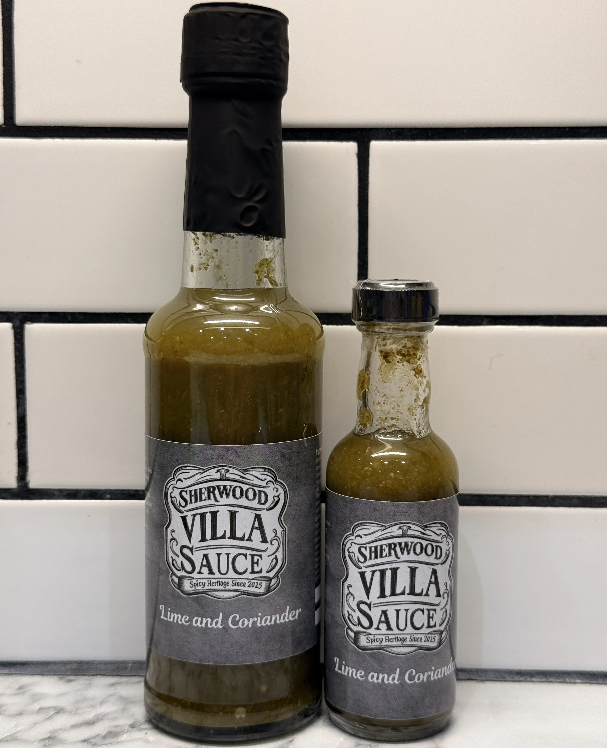 Two bottles of Sherwood Villa Sauce lime and coriander on a marble surface in front of a white and black tiled wall.