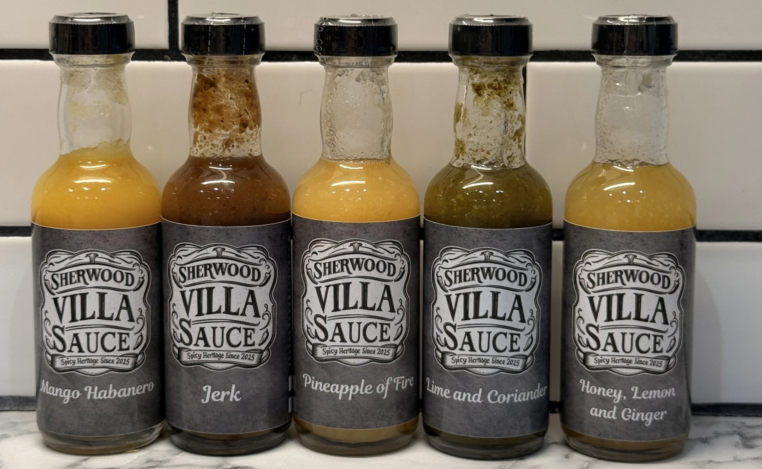 Five bottles of Sherwood Villa Sauce with different flavors: Mango Habanero, Jerk, Pineapple of Fire, Lime and Coriander, Honey Lemon and Ginger, placed on a marble surface against a tiled wall.