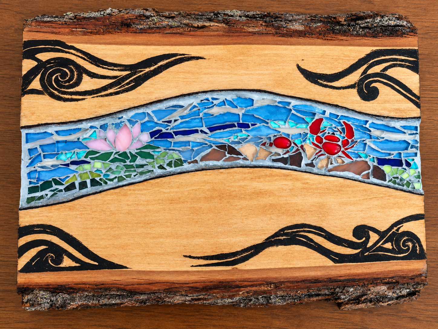 A decorative wooden piece with a mosaic of a landscape scene. The scene includes a pink lotus flower on the left, a red crab on the right, a body of water with blue glass tiles, and black swirling patterns above and below. The wood has a natural bark edge at the top and bottom.