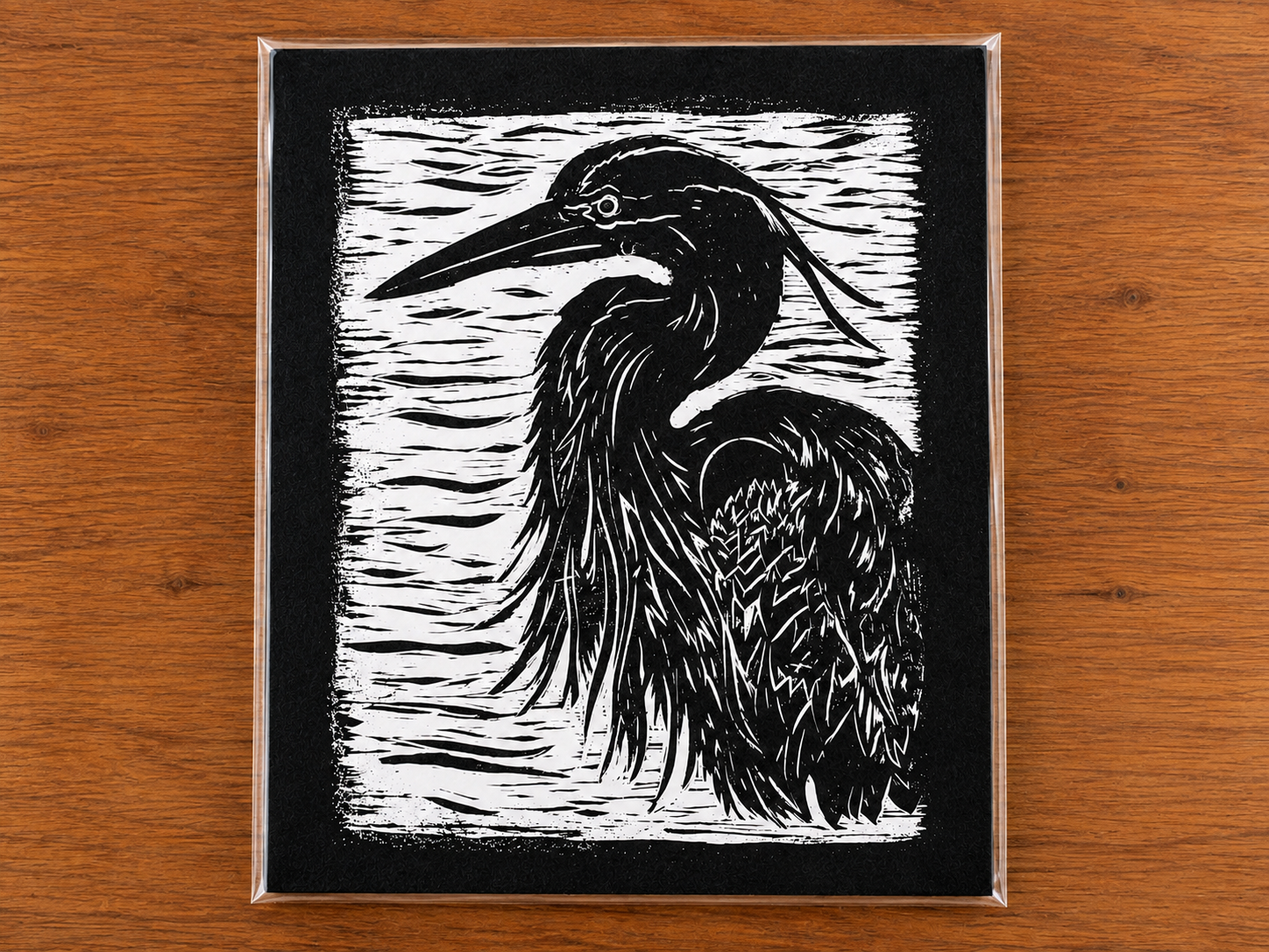 Silhouette painting of a heron bird on a textured black and white background, framed in a clear case, placed on a wooden surface.