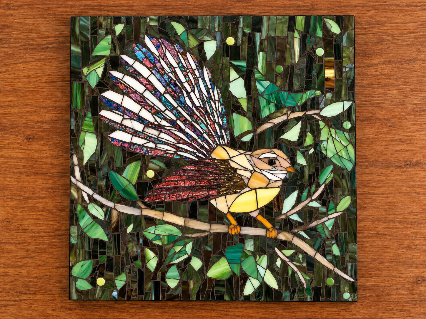 Colorful stained glass mosaic of a bird with vibrant wings perched on a branch amid green leaves.