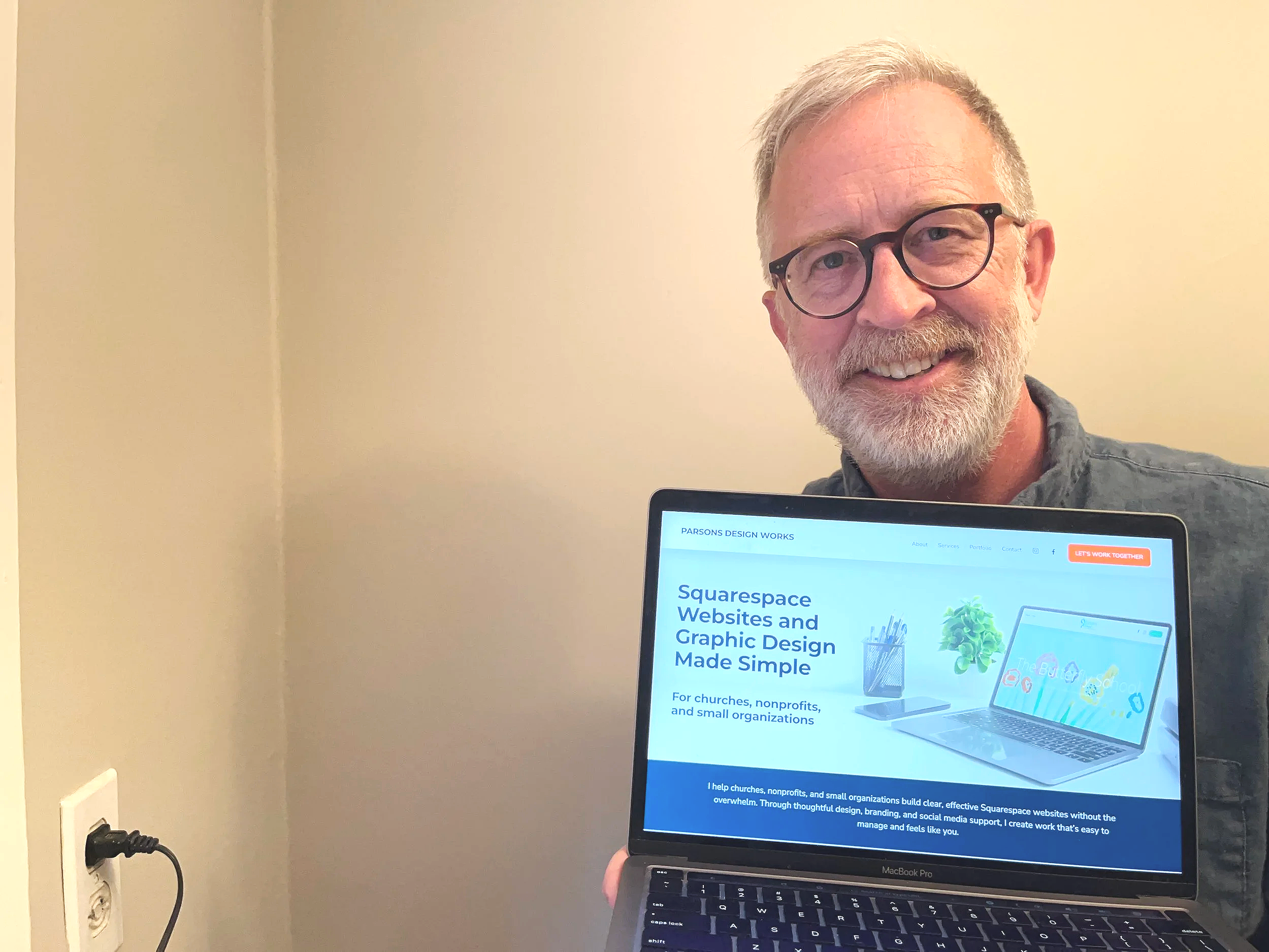 A smiling man with glasses and a beard holding a laptop displaying a website for Squarespace, which offers website and graphic design services for small organizations.