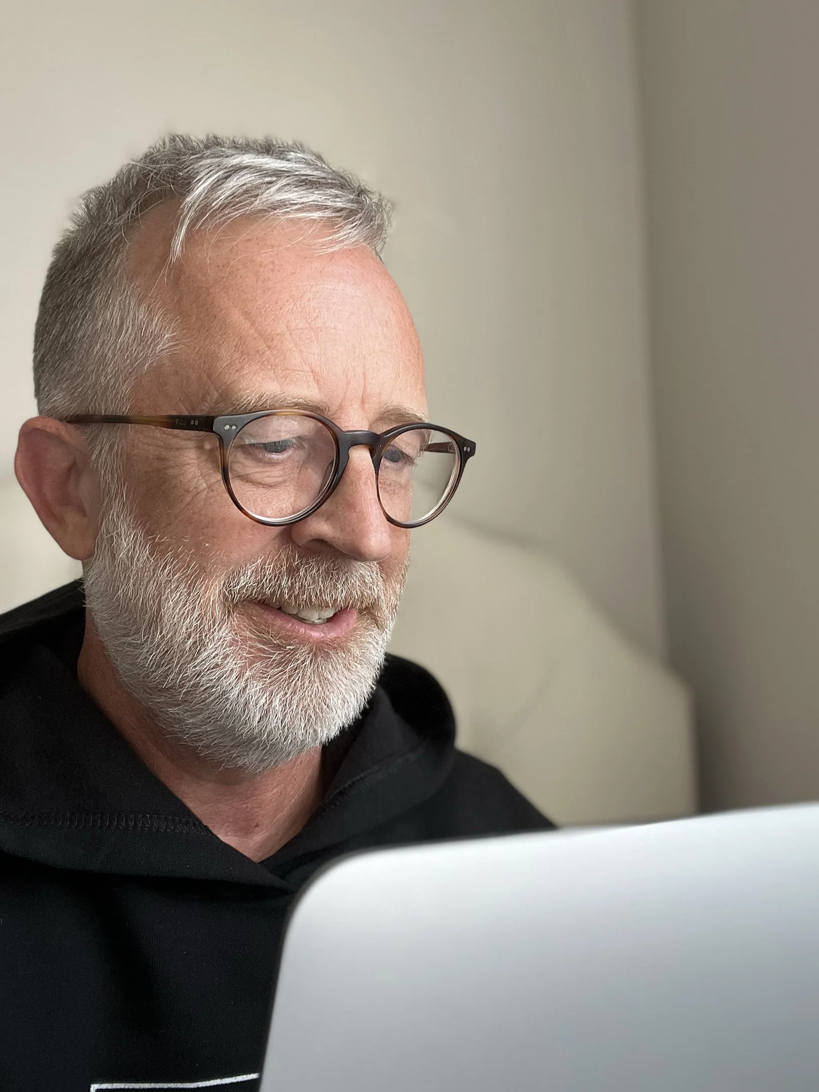 Close-up of an older man with gray hair, beard, and glasses, working on a computer in a neutral-colored room.