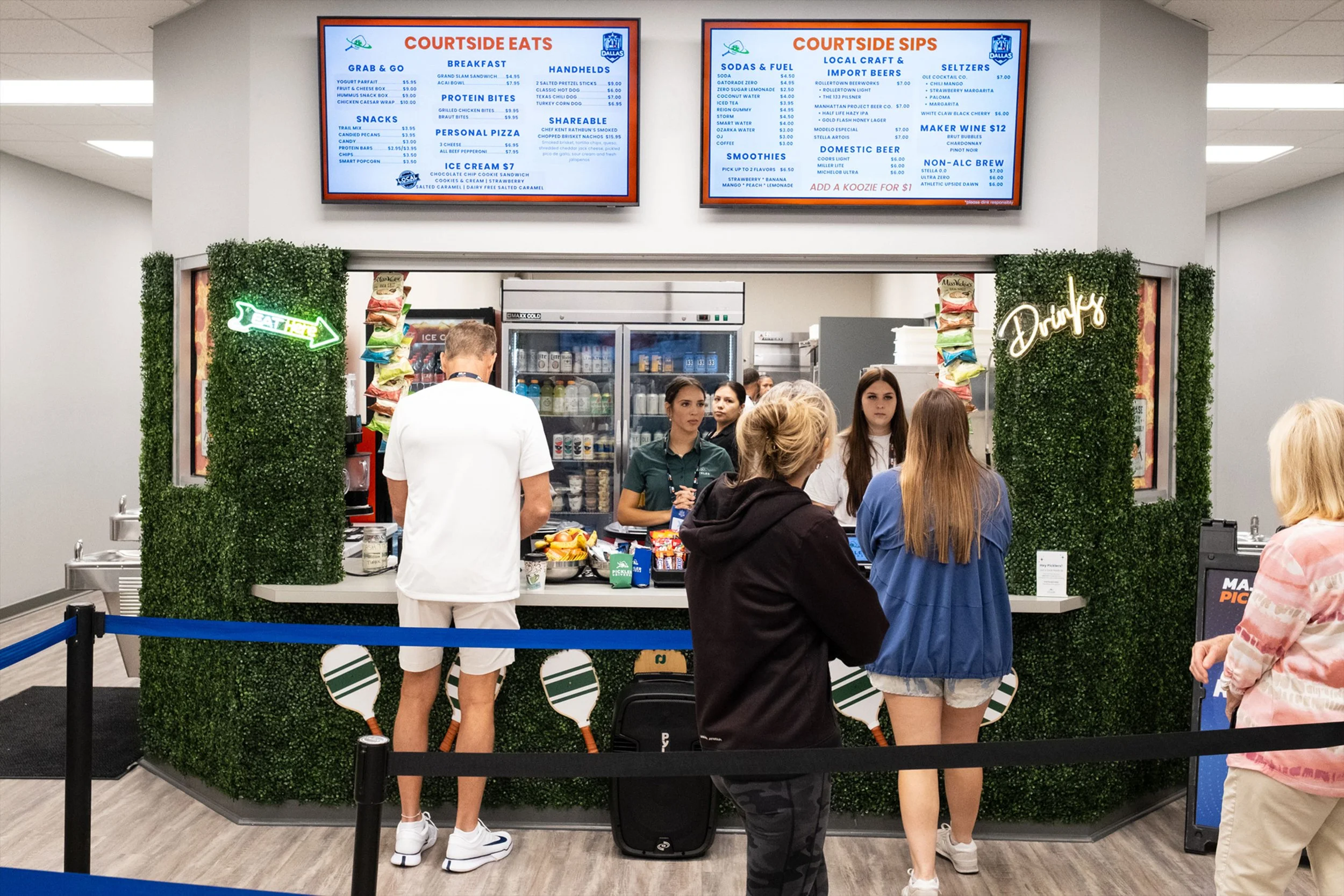 People standing in line at a food kiosk called Courtside Eats, which offers breakfast, snacks, personal pizza, ice cream, and drinks, with a digital menu board displaying food and beverage options above.