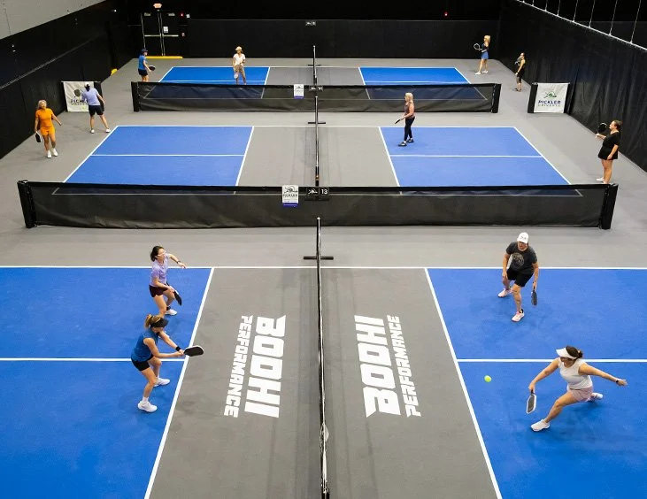Indoor pickleball courts with players engaged in game, wearing athletic clothing, surrounded by black barriers with white logos, and a gray floor.