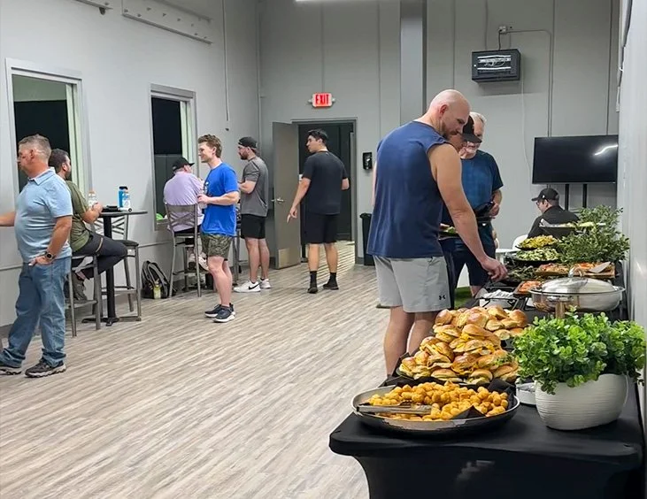 People at a casual indoor gathering with a food buffet, including sandwiches, chips, and various dishes, in a modern room with gray walls and wood flooring.
