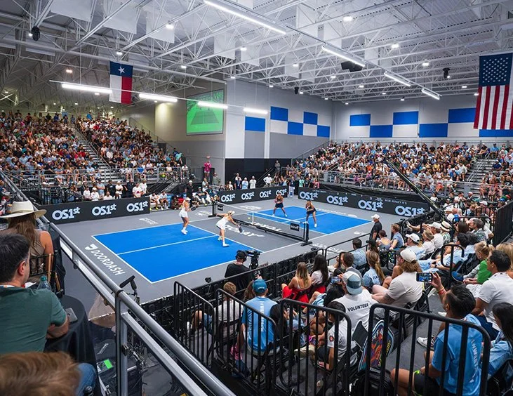 Indoor tennis match with two teams playing on a blue court, surrounded by a large crowd in the stands and spectators seated near the court.
