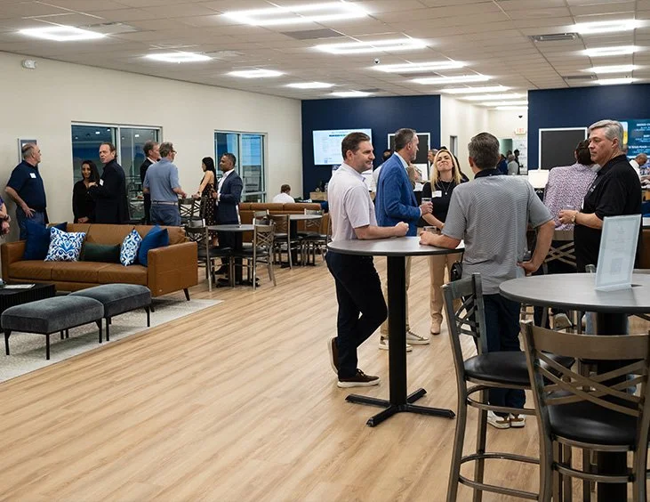 People socializing and networking at a professional event in a modern, well-lit office or conference space with wooden floors, lounge seating, and multiple screens on the walls.