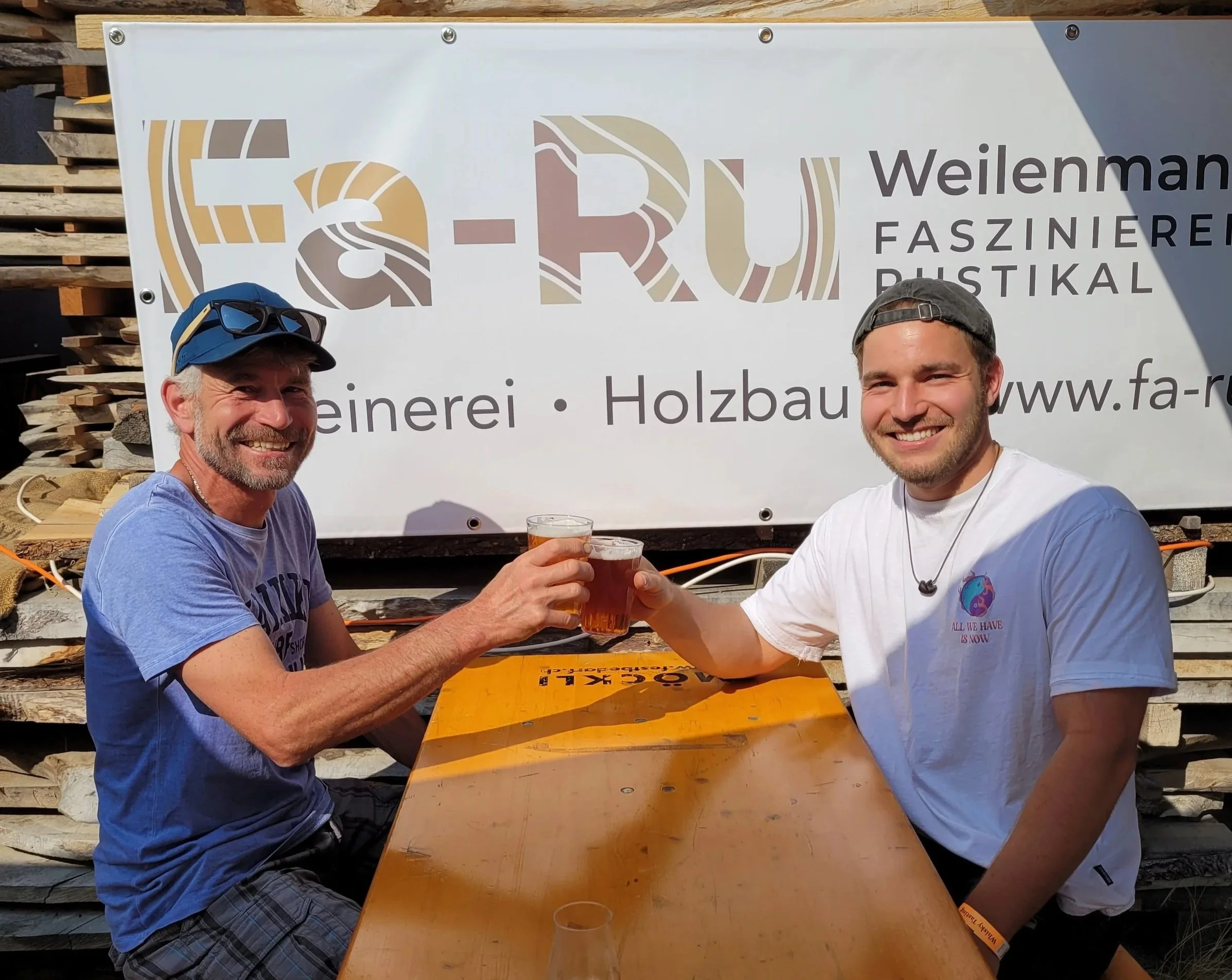 Zwei Männer sitzen an einem Holztisch im Freien und stoßen mit Biergläsern an, vor einem Holzzaun. Hinter ihnen ein großes Banner mit dem Text Fa-Ru Weilenmann Faszinierend Rustikal