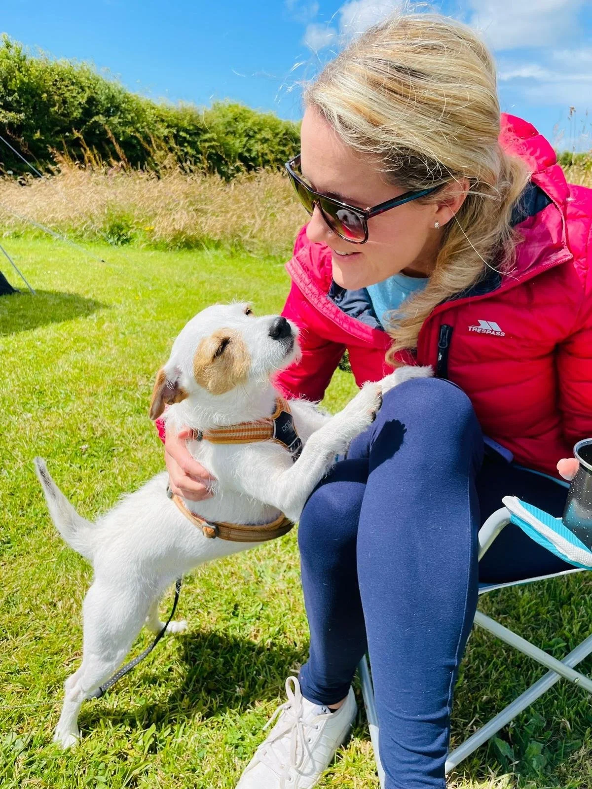 A woman in sunglasses and a pink jacket is smiling and holding a small white dog with a brown patch on its face, outdoors on a sunny day with green grass and blue sky.