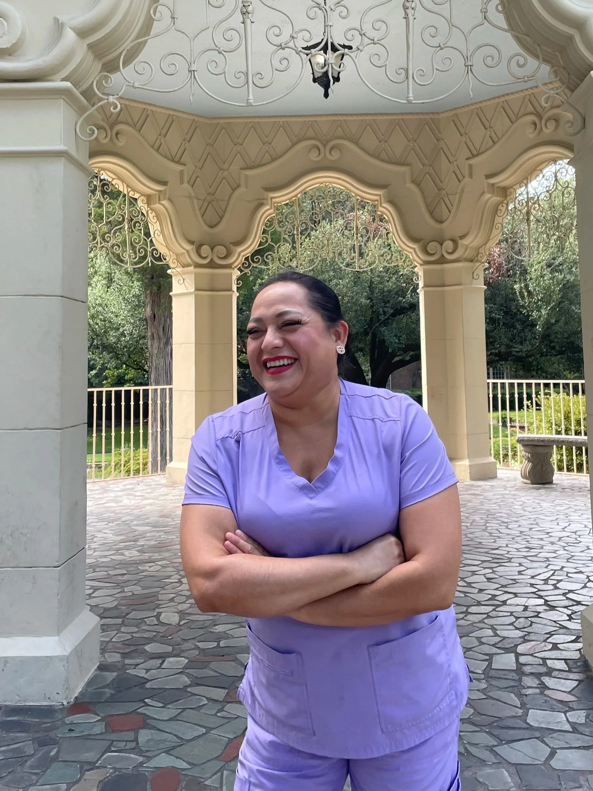 A woman in light purple medical scrubs standing outdoors under a decorative gazebo, smiling and crossing her arms.