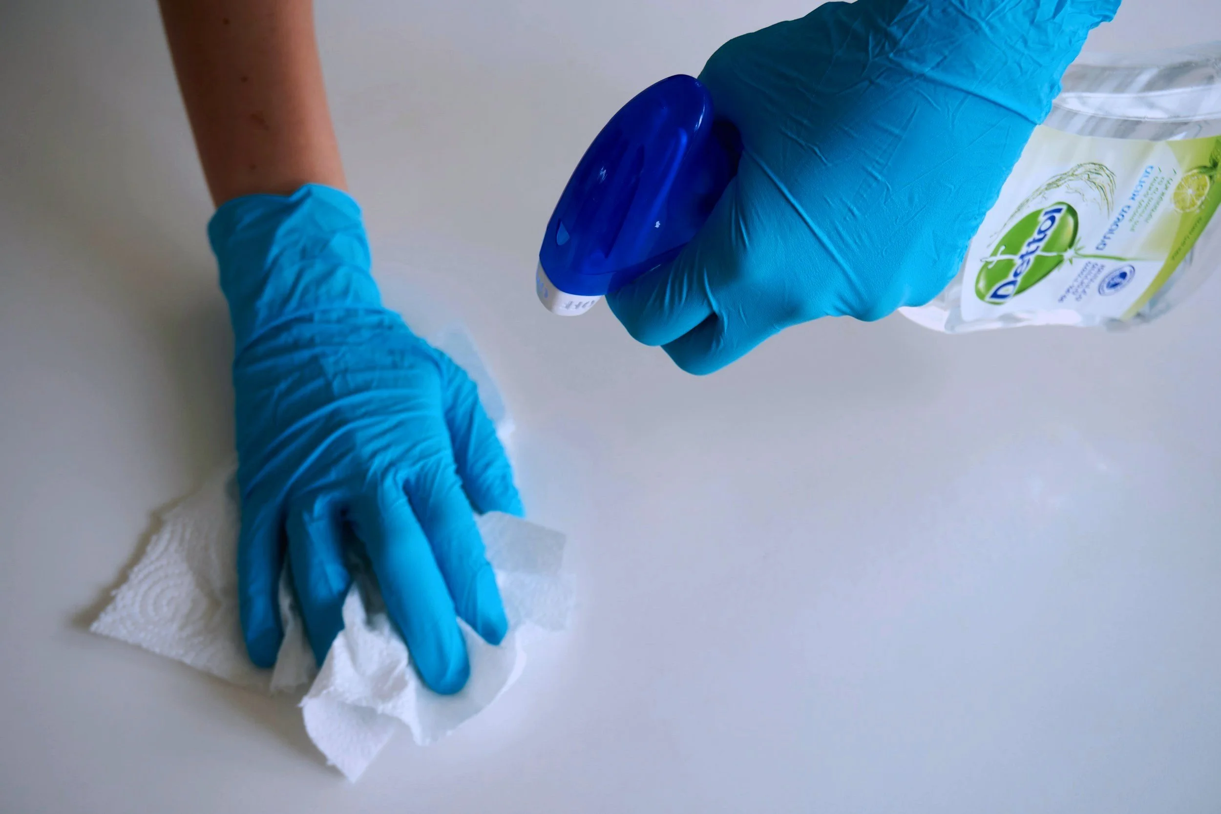 Person cleaning a countertop with a paper towel and wearing blue gloves, holding a bottle of eco-friendly cleaning spray.