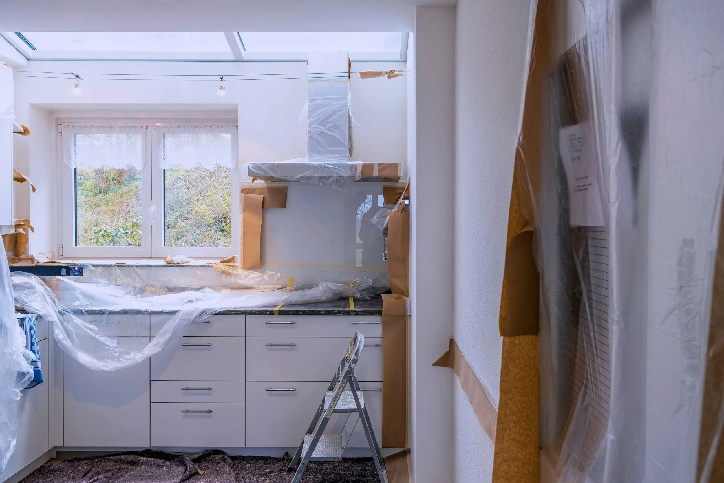 Kitchen under renovation with plastic sheeting, a step ladder, and wrapped fixtures, with a large window showing outdoors.