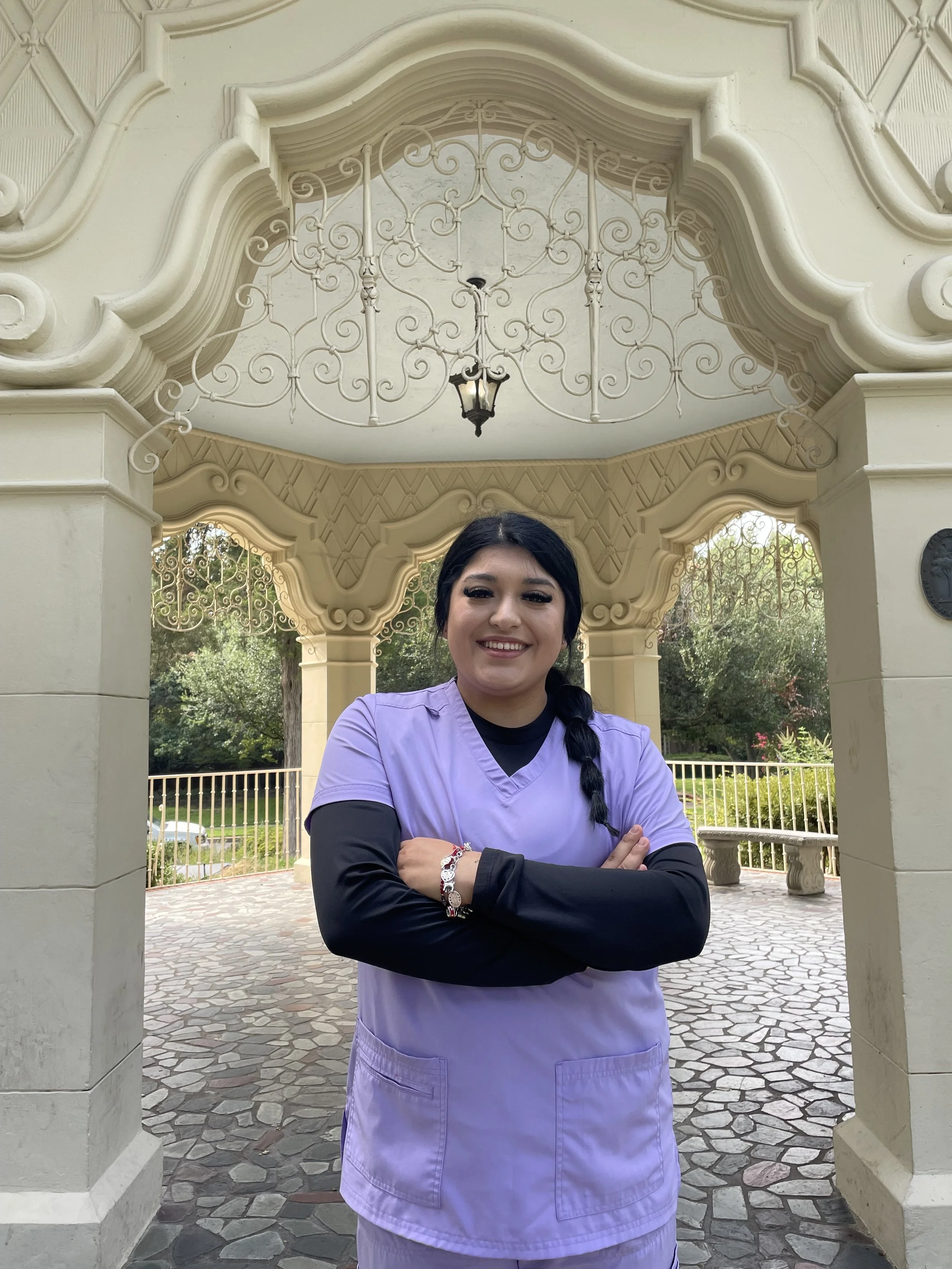 Smiling woman in purple scrubs with black sleeves standing outdoors under ornate beige archway with ironwork and lantern, crossed arms.