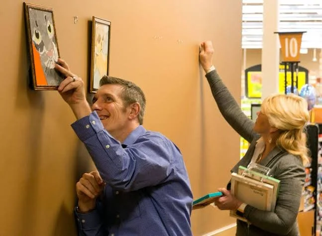 A man and woman in a store laughing and pretending to steal framed pictures, with the woman holding a clipboard and books.
